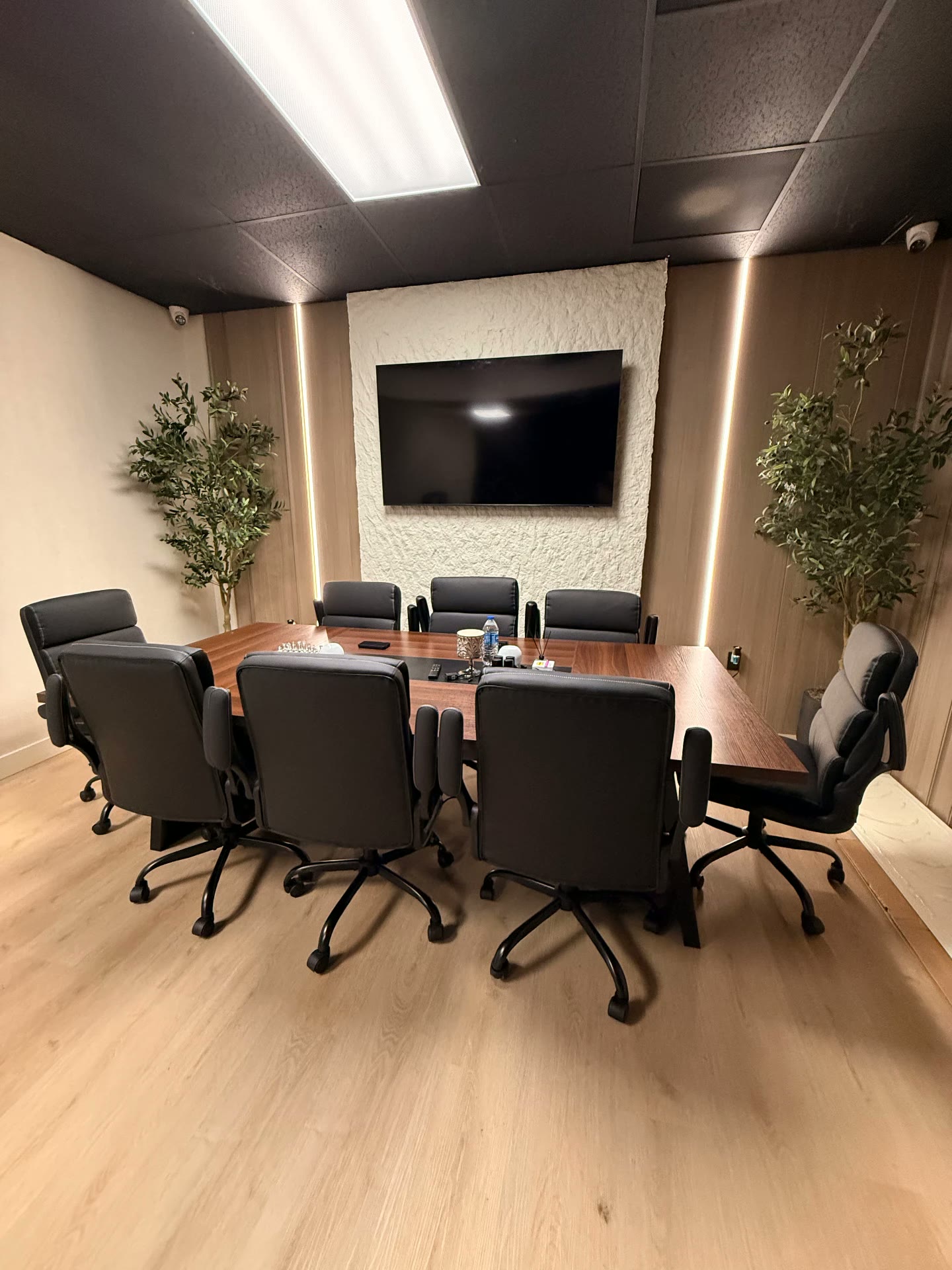 Sala de juntas corporativa con mesa de madera nogal, ocho sillas ejecutivas negras, paneles de madera con iluminación LED vertical, muro texturizado con pantalla y olivos decorativos