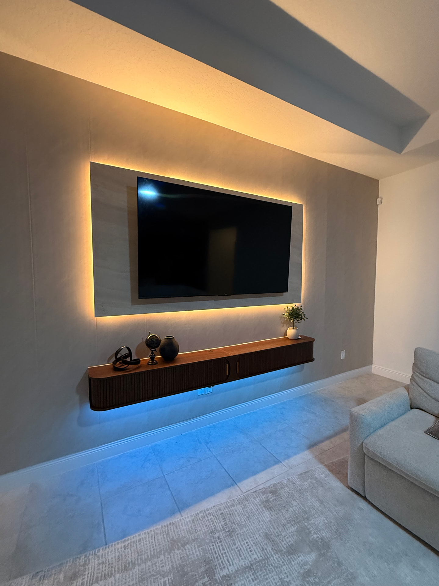 Muro de TV en microcemento con marco de porcelanato retroiluminado en luz cálida, mueble flotante acanalado de nogal con luz LED azul y sillón gris claro