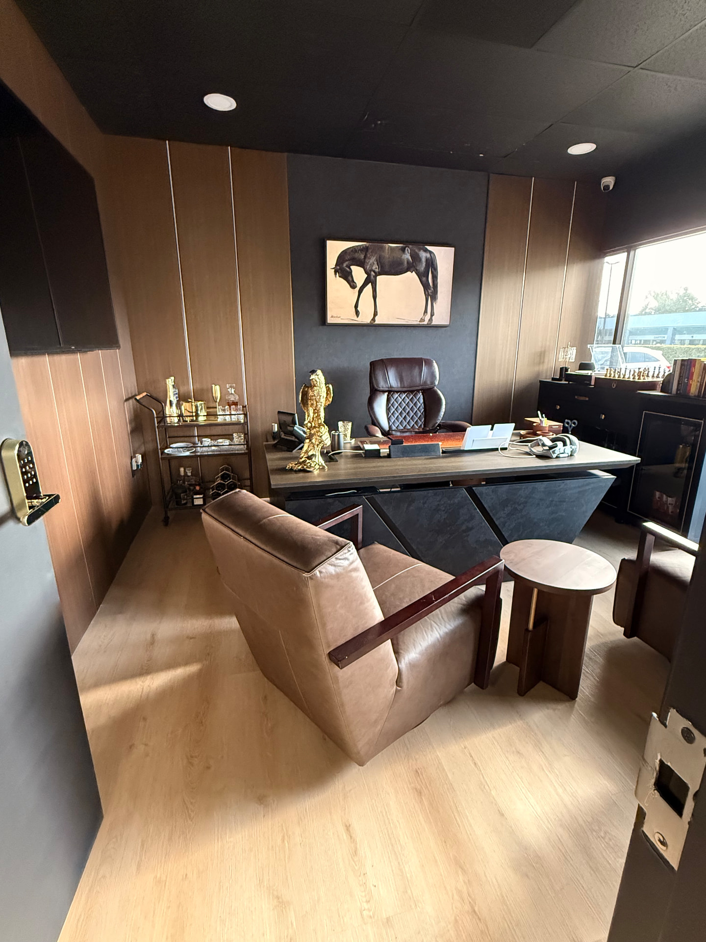 Oficina ejecutiva con paneles de madera roble, techo negro, escritorio flotante gris, sillón de cuero, carrito de bar dorado y cuadro de caballo sobre muro texturizado oscuro