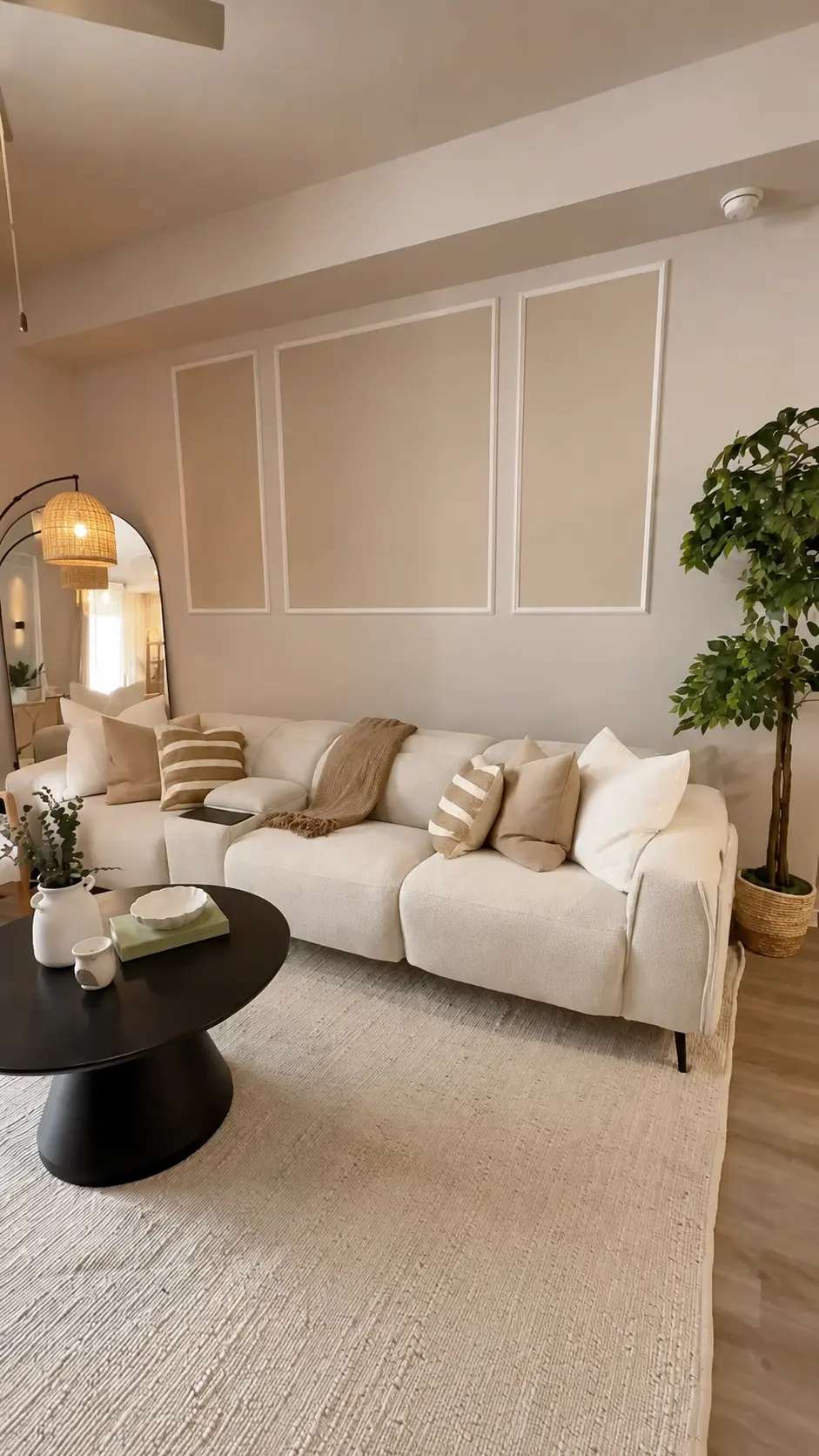 Sala residencial en tonos crema y beige con sofá modular blanco, cojines de lino y rayas, molduras decorativas en la pared, alfombra de yute, mesa de centro negra redonda y planta de interior